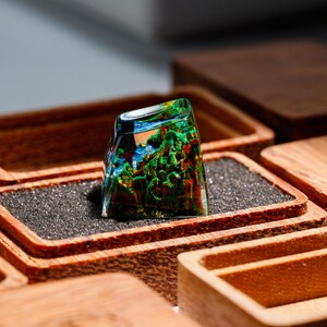 May include: A clear, rectangular resin keycap with a miniature landscape scene inside, featuring green trees, blue water, and a colorful sky. The keycap is set in a wooden box with a dark gray interior.