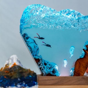 May include: A blue resin sculpture of an underwater scene with two divers and jellyfish. The resin is clear and has a wave-like texture at the top. The sculpture is mounted on a wooden base.