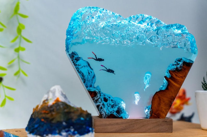 May include: A blue resin sculpture of an underwater scene with two divers and jellyfish. The resin is clear and has a wave-like texture at the top. The sculpture is mounted on a wooden base.