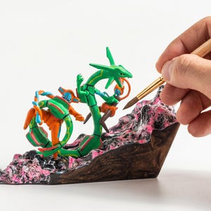May include: A green serpentine dragon figure and an orange humanoid figure with blue accents positioned on a textured, rocky base with black and pink details. The figures are sculpted in a dynamic pose, with the dragon coiled around the base.