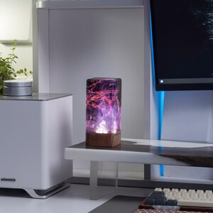 May include: A rectangular resin lamp with a wooden base. The lamp's interior features a swirling pattern of purple and red hues, illuminated from within. It sits on a white table near a white speaker and a computer monitor.