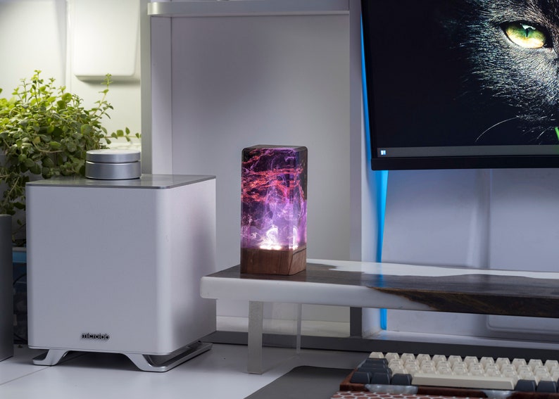 May include: A rectangular resin lamp with a wooden base. The lamp's interior features a swirling pattern of purple and red hues, illuminated from within. It sits on a white table near a white speaker and a computer monitor.
