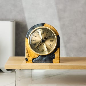 May include: A decorative clock with a gold-colored face and Roman numerals. The clock is set in a wooden frame with a unique pattern of black and gold tones. The clock sits on a wooden shelf.