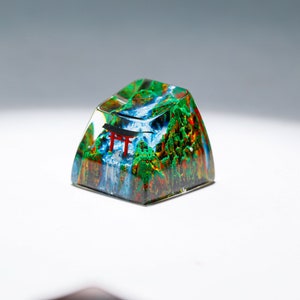 May include: A custom keycap featuring a miniature landscape with a waterfall, trees, and a red torii gate. The keycap is encased in clear resin, creating a 3D effect. The colors include green, blue, red, and brown.