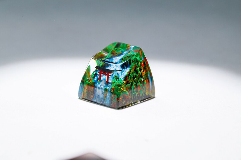 May include: A custom keycap featuring a miniature landscape with a waterfall, trees, and a red torii gate. The keycap is encased in clear resin, creating a 3D effect. The colors include green, blue, red, and brown.