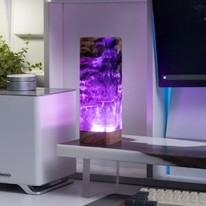 May include: A rectangular table lamp with a wooden base and a translucent purple resin body. The lamp is lit from within, creating a glowing effect. The lamp is on a white desk with a computer and other items.