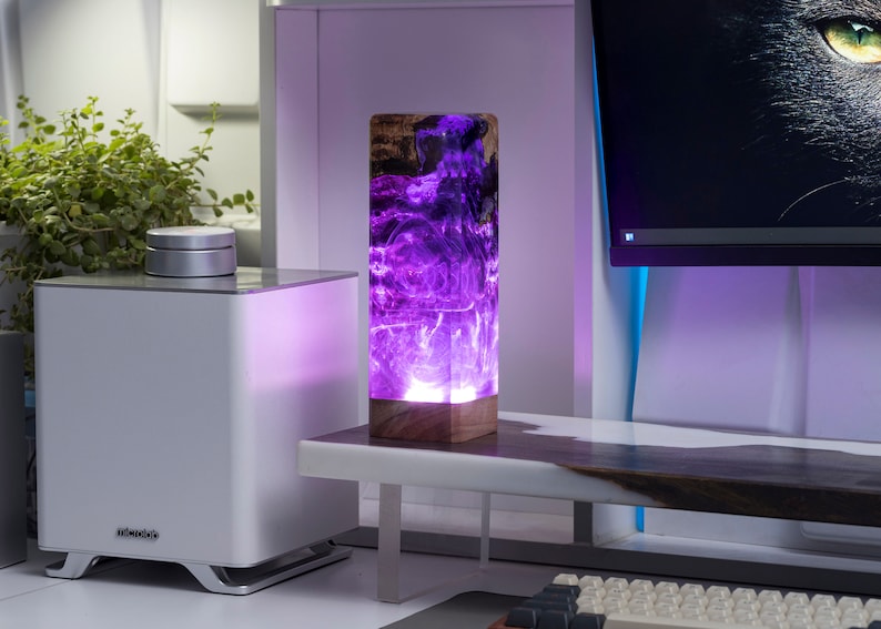 May include: A rectangular table lamp with a wooden base and a translucent purple resin body. The lamp is lit from within, creating a glowing effect. The lamp is on a white desk with a computer and other items.