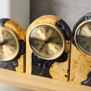 May include: Three decorative mantel clocks with gold-tone faces and Roman numeral hour markers. The clocks have a unique design, featuring a combination of light wood and black resin. The clock faces are encased in a gold-colored metal frame.