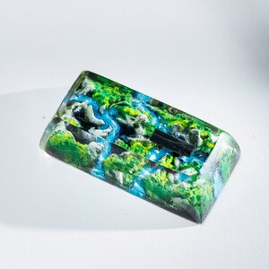 May include: A rectangular resin art piece featuring a miniature landscape with a river, green hills, and white rock formations. The artwork is encased in clear resin, creating a 3D effect. The color palette includes shades of green, blue, and white.