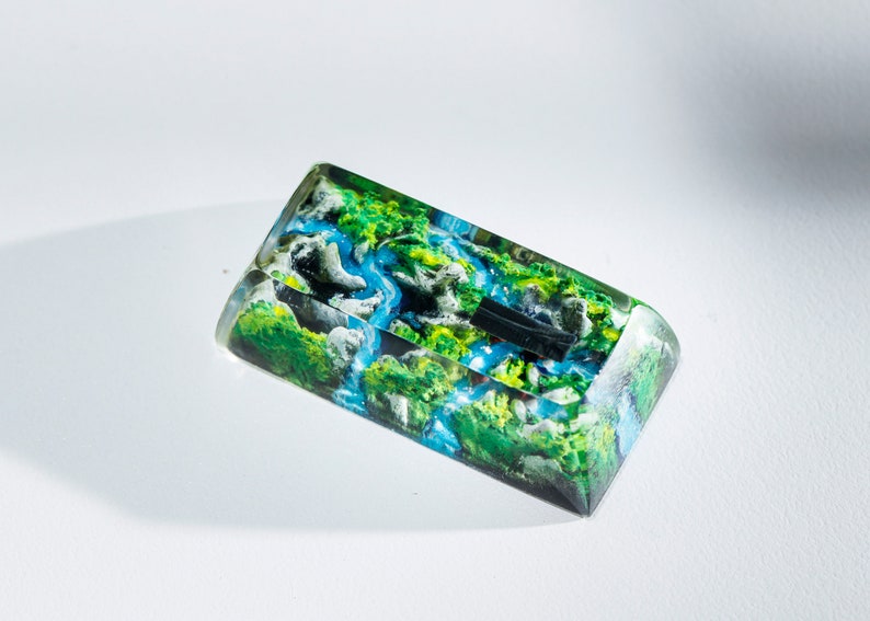 May include: A rectangular resin art piece featuring a miniature landscape with a river, green hills, and white rock formations. The artwork is encased in clear resin, creating a 3D effect. The color palette includes shades of green, blue, and white.