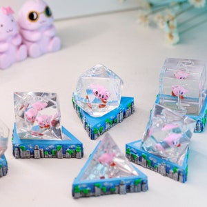 May include: Clear resin sculptures with pink cartoon characters inside geometric shapes. Each sits on a blue base with a cityscape design. Shapes include cubes, pyramids, and a multi-sided die. The sculptures are decorative.