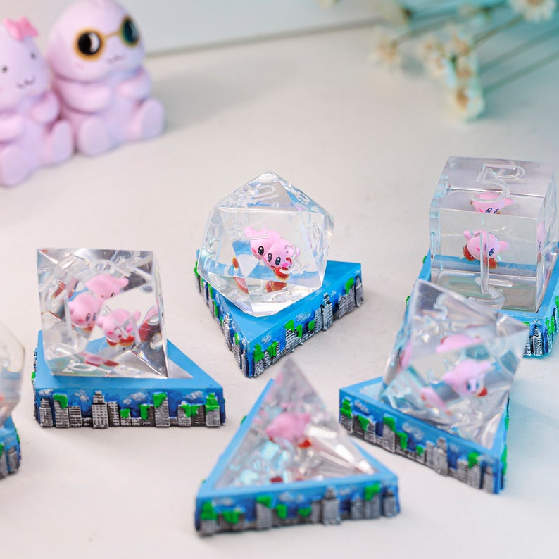 May include: Clear resin sculptures with pink cartoon characters inside geometric shapes. Each sits on a blue base with a cityscape design. Shapes include cubes, pyramids, and a multi-sided die. The sculptures are decorative.