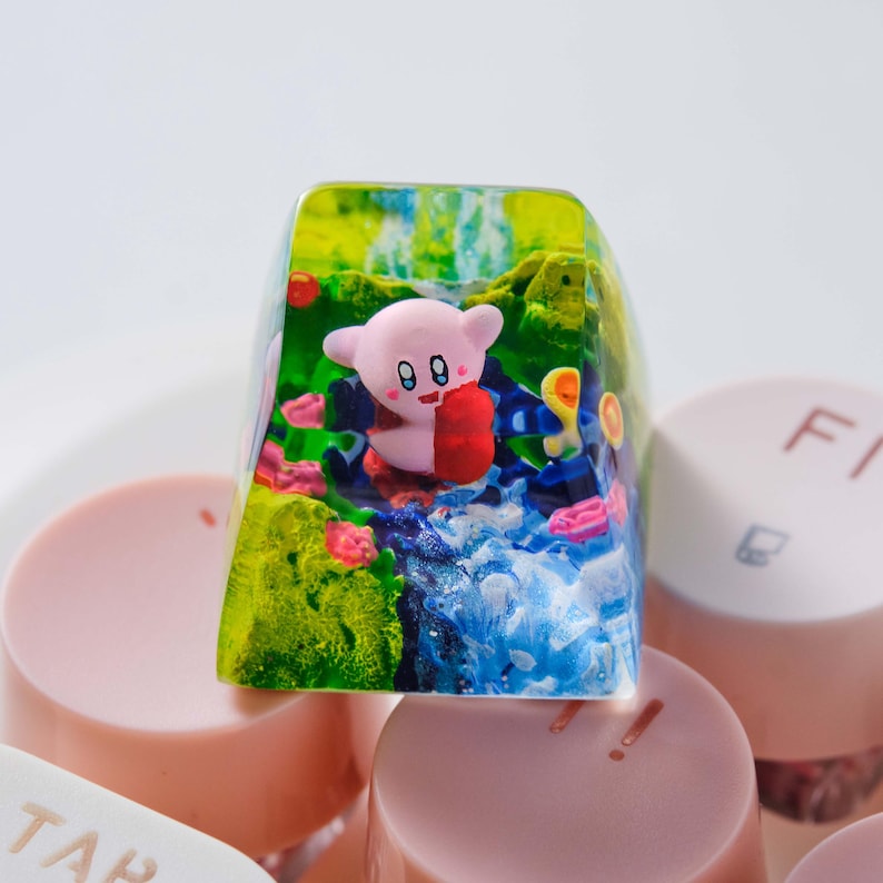 May include: A custom resin mechanical keyboard keycap featuring a miniature pink Kirby character holding a red object, set within a translucent green and blue landscape with textured details, positioned among light pink keyboard keys.