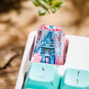 Puede incluir: Una tecla de teclado personalizada con un dise&ntilde;o colorido. La tecla presenta una escena de templo japon&eacute;s en miniatura con tonos rojos, azules y rosas. La tecla est&aacute; colocada en una bandeja de teclado blanca, con teclas azul claro visibles.