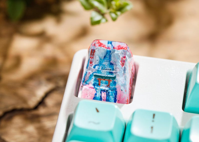 Puede incluir: Una tecla de teclado personalizada con un dise&ntilde;o colorido. La tecla presenta una escena de templo japon&eacute;s en miniatura con tonos rojos, azules y rosas. La tecla est&aacute; colocada en una bandeja de teclado blanca, con teclas azul claro visibles.