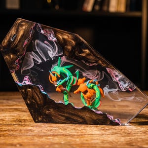 May include: A trapezoidal resin display piece featuring a green and orange serpentine creature figurine encased within a clear center. The surrounding frame is made of dark, swirling metallic brown resin with pink and white accents and wispy smoke.