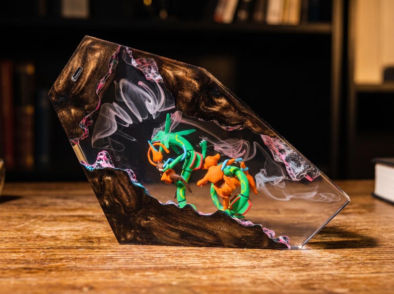 May include: A trapezoidal resin display piece featuring a green and orange serpentine creature figurine encased within a clear center. The surrounding frame is made of dark, swirling metallic brown resin with pink and white accents and wispy smoke.