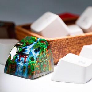May include: A custom keycap with a detailed landscape design featuring a waterfall, trees, and a red torii gate. The keycap is surrounded by other white keycaps and a wooden container.