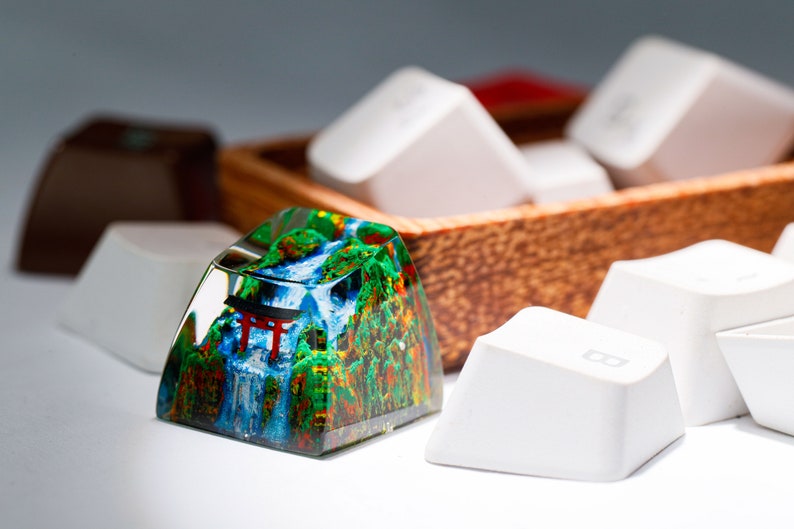 May include: A custom keycap with a detailed landscape design featuring a waterfall, trees, and a red torii gate. The keycap is surrounded by other white keycaps and a wooden container.
