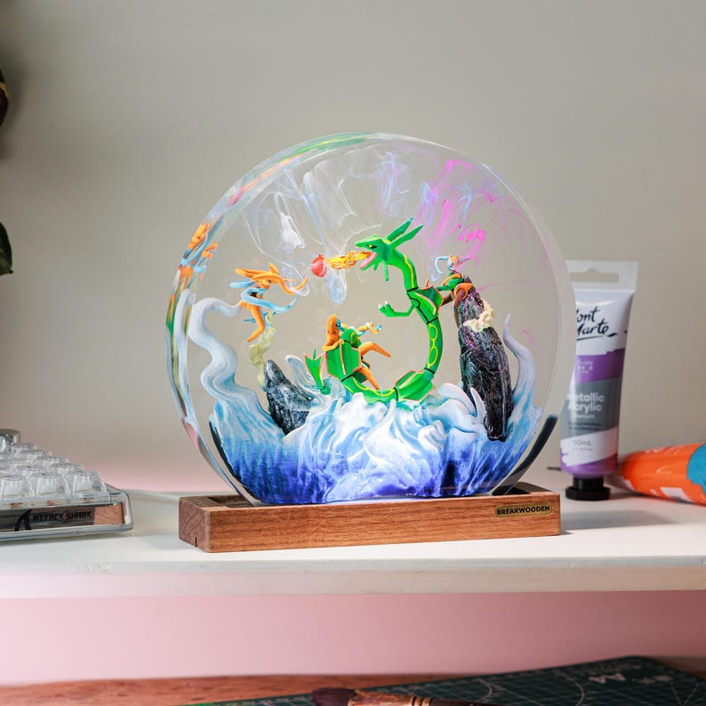 May include: A circular resin diorama featuring a green serpentine creature and orange figures amidst swirling white and blue clouds, set on a rectangular wooden base with a small plaque labeled BREAKWOODEN, creating a detailed scene.
