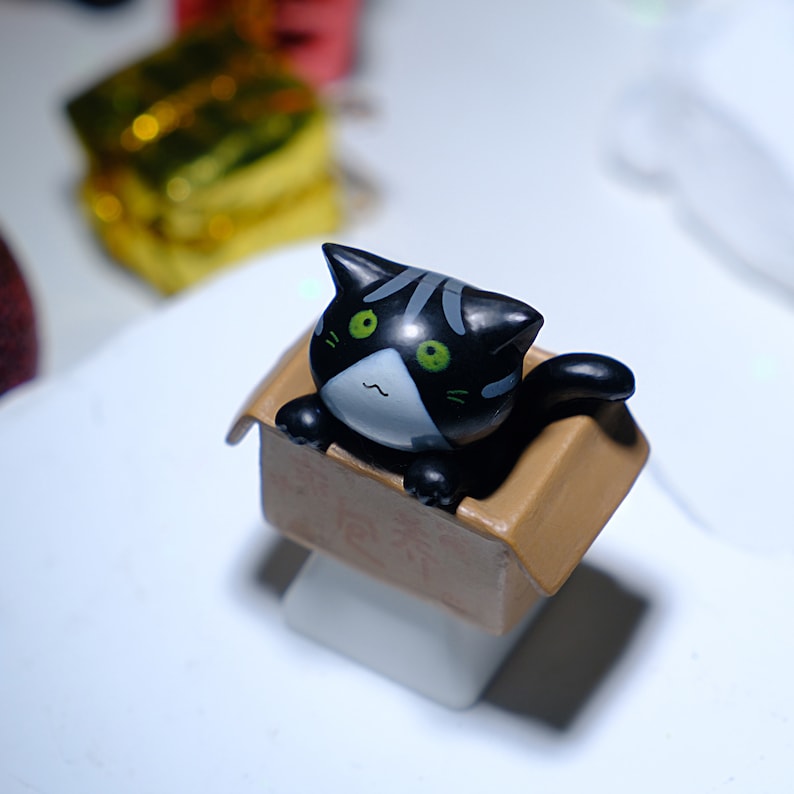 May include: A black cat figurine with green eyes and gray stripes peeks out of a small cardboard box. The box is tan and sits on a white surface. The cat has a white face and a curved mouth.