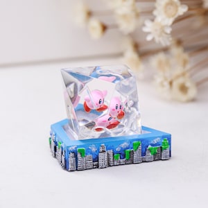 May include: A clear resin sculpture in a geometric shape, with two pink cartoon characters inside. The sculpture is set on a blue base with a cityscape design. The characters are encased in the clear resin.