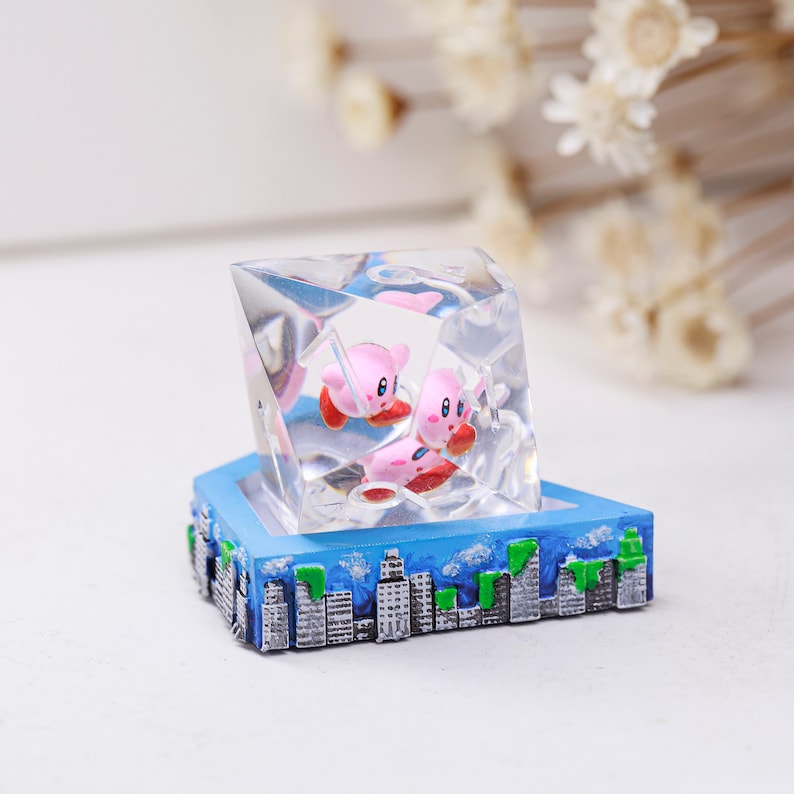 May include: A clear resin sculpture in a geometric shape, with two pink cartoon characters inside. The sculpture is set on a blue base with a cityscape design. The characters are encased in the clear resin.