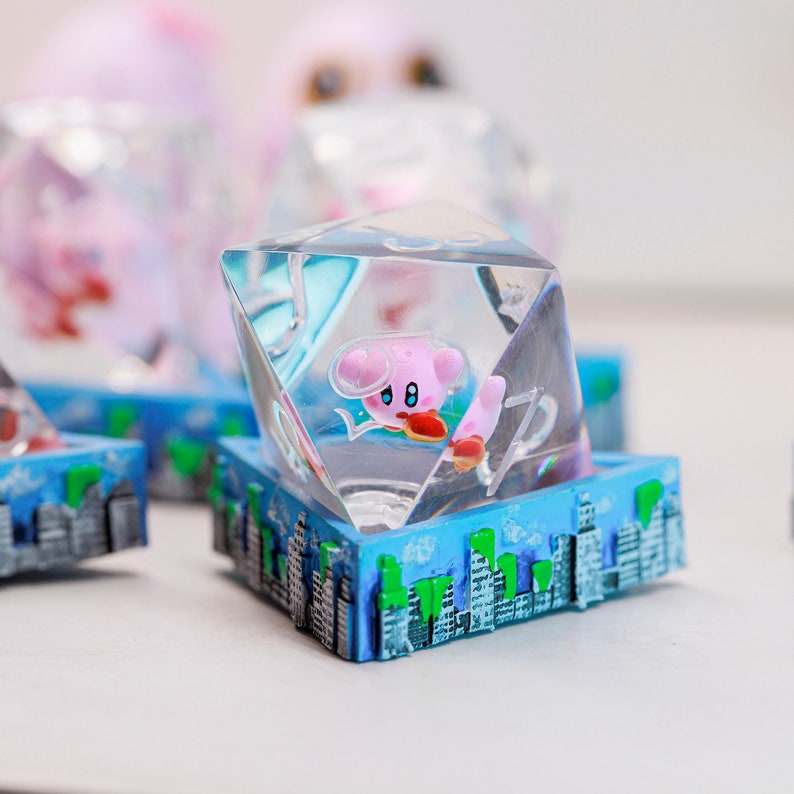 May include: A clear, multi-faceted resin die with a pink cartoon character in a space helmet. The die sits on a blue base with a cityscape design. Other similar dice are visible in the background.