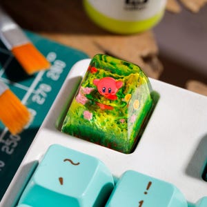 May include: A custom resin mechanical keyboard keycap featuring a small pink character embedded within a translucent green and yellow landscape design, installed on a white keyboard with light blue keys.