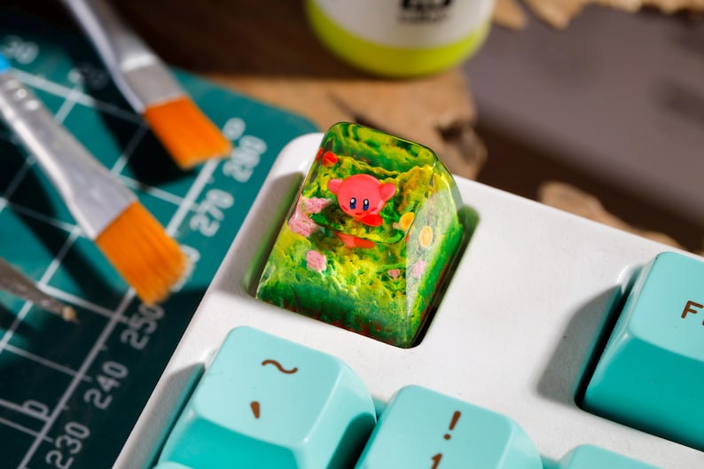 May include: A custom resin mechanical keyboard keycap featuring a small pink character embedded within a translucent green and yellow landscape design, installed on a white keyboard with light blue keys.