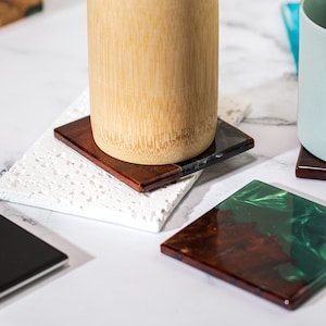 May include: Square coasters in various designs. One coaster is a dark wood with a clear resin edge, another is a white speckled stone, and a third is a dark wood with green resin. A bamboo cup sits on one coaster.