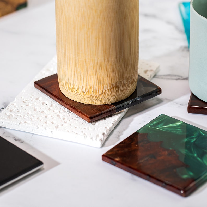 May include: Square coasters in various designs. One coaster is a dark wood with a clear resin edge, another is a white speckled stone, and a third is a dark wood with green resin. A bamboo cup sits on one coaster.