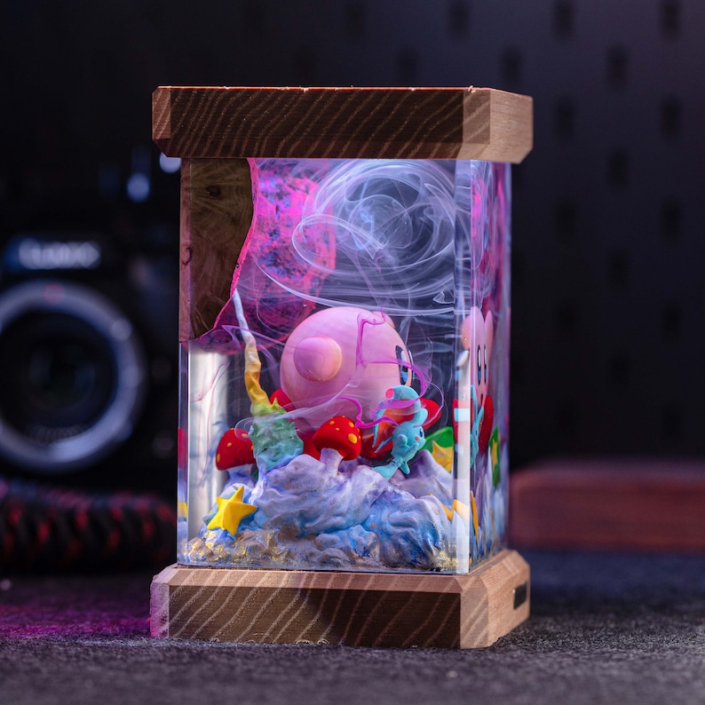 May include: A rectangular resin lamp featuring a pink spherical character inside, surrounded by miniature red mushrooms, yellow stars, and blue cloud-like formations. The clear resin contains swirling white smoke effects, encased between two horizontal wooden blocks.