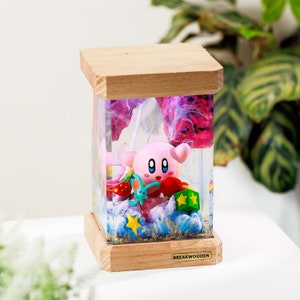 May include: A rectangular resin lamp with a light wood base and top. The clear resin body contains a pink Kirby figure, a small blue creature, red mushrooms, a green star block, and a yellow star. The base features a label reading BREAKWOODEN.