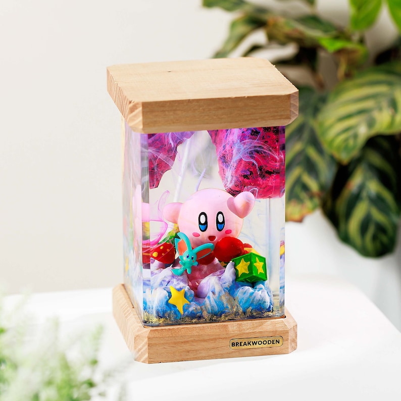 May include: A rectangular resin lamp with a light wood base and top. The clear resin body contains a pink Kirby figure, a small blue creature, red mushrooms, a green star block, and a yellow star. The base features a label reading BREAKWOODEN.