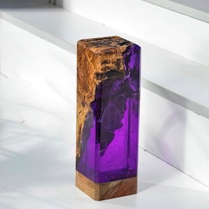 May include: A rectangular wooden block with a striking design. The top and base are natural wood, while the center is a vibrant purple resin. The wood grain contrasts with the smooth resin, creating a unique decorative piece.