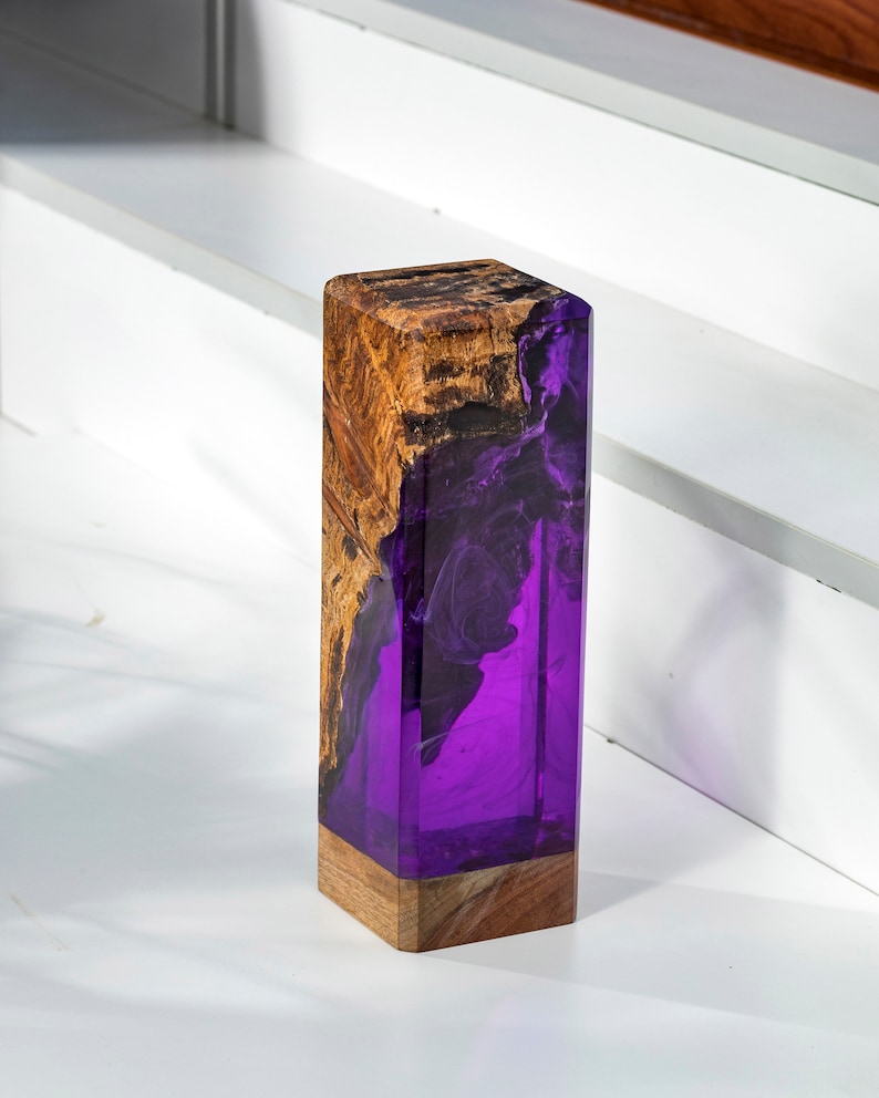 May include: A rectangular wooden block with a striking design. The top and base are natural wood, while the center is a vibrant purple resin. The wood grain contrasts with the smooth resin, creating a unique decorative piece.