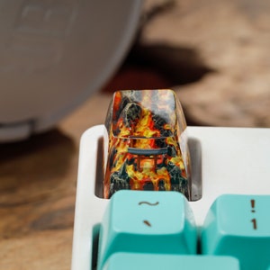 Op de afbeelding: Close-up van een aangepaste keycap met een vurig, lava-achtig ontwerp in rood, oranje en geel. De keycap is ge&iuml;nstalleerd op een wit toetsenbord met lichtblauwe toetsen. De keycap heeft een unieke, artistieke uitstraling.