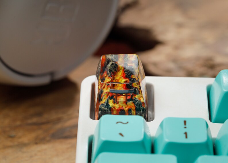 Op de afbeelding: Close-up van een aangepaste keycap met een vurig, lava-achtig ontwerp in rood, oranje en geel. De keycap is ge&iuml;nstalleerd op een wit toetsenbord met lichtblauwe toetsen. De keycap heeft een unieke, artistieke uitstraling.