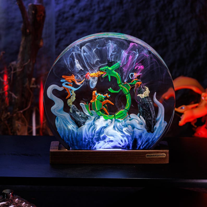 May include: A circular resin lamp featuring a green serpentine creature and orange figures amidst swirling blue and white smoke effects. The piece is mounted on a rectangular wooden base with the text BREAKWOODEN printed on a small metal plaque.