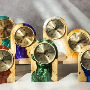 May include: A collection of wooden mantel clocks with gold-tone clock faces and Roman numerals. The clocks have a variety of colorful resin accents in shades of green, blue, purple, orange, and red. The clocks are displayed on a light-colored surface.