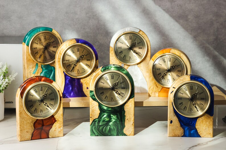 May include: A collection of wooden mantel clocks with gold-tone clock faces and Roman numerals. The clocks have a variety of colorful resin accents in shades of green, blue, purple, orange, and red. The clocks are displayed on a light-colored surface.