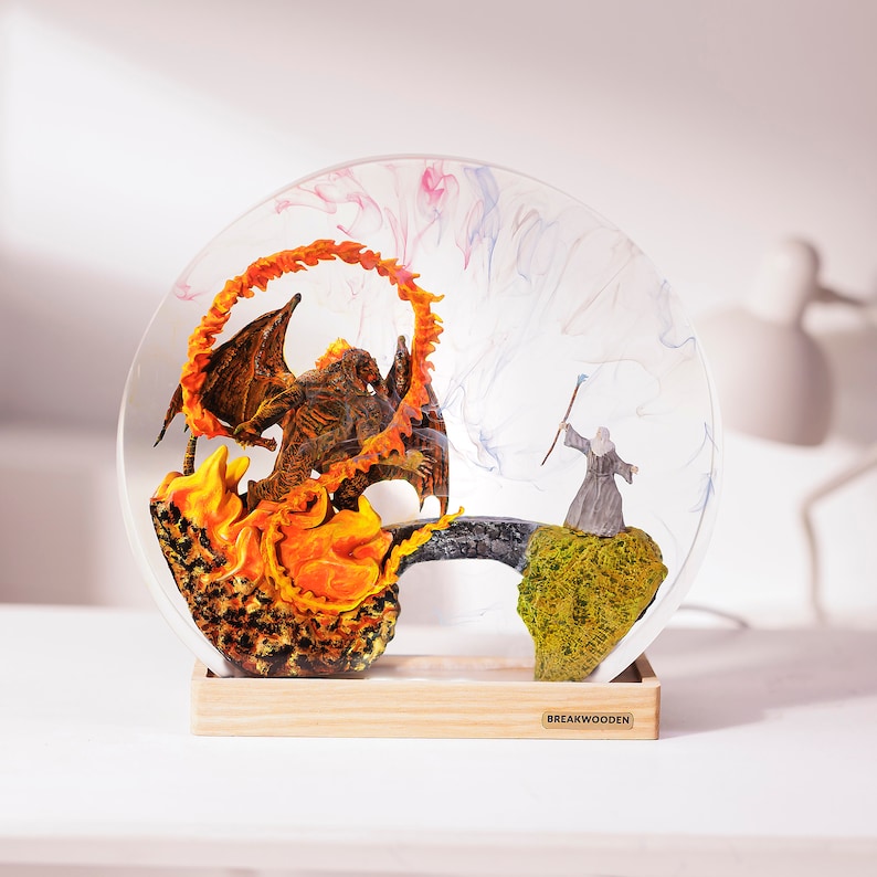 May include: A decorative resin sculpture featuring a fiery demon and a wizard on a bridge, set within a clear, circular frame. The base is made of light-colored wood. The sculpture has a fantasy theme.