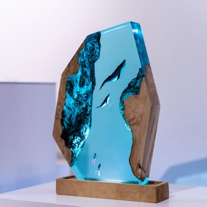 May include: A unique, illuminated resin sculpture featuring a marine scene. The artwork showcases a light blue, transparent resin with embedded whale figures and divers. The sculpture is framed by natural wood edges and sits on a wooden base, creating a striking contrast.