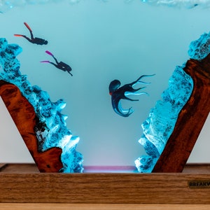 May include: A triangular resin sculpture with a blue and white ocean scene. Two divers are swimming in the water, and an octopus is in the middle of the scene. The sculpture is lit from below with a pink light. The sculpture is on a wooden base with the text "BREAKWOODEN" on the bottom.