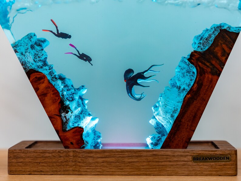 May include: A triangular resin sculpture with a blue and white ocean scene. Two divers are swimming in the water, and an octopus is in the middle of the scene. The sculpture is lit from below with a pink light. The sculpture is on a wooden base with the text "BREAKWOODEN" on the bottom.