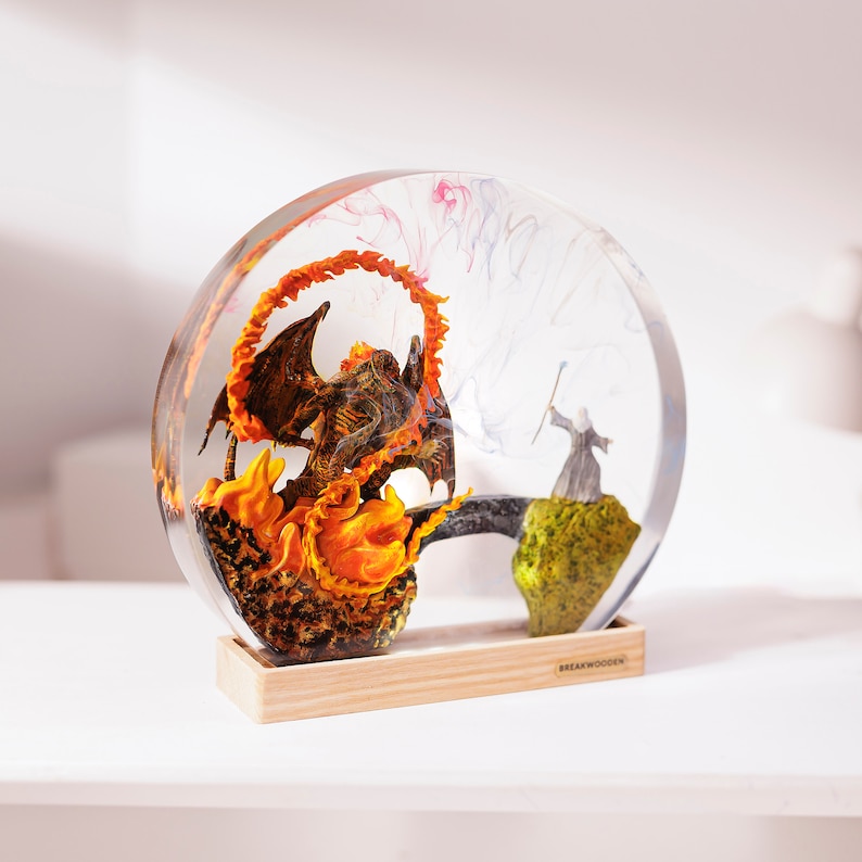 May include: A clear resin sphere encases a fantasy scene. A fiery dragon and a wizard stand on a bridge and a mossy hill. The base is made of light-colored wood. The dragon is surrounded by orange flames.