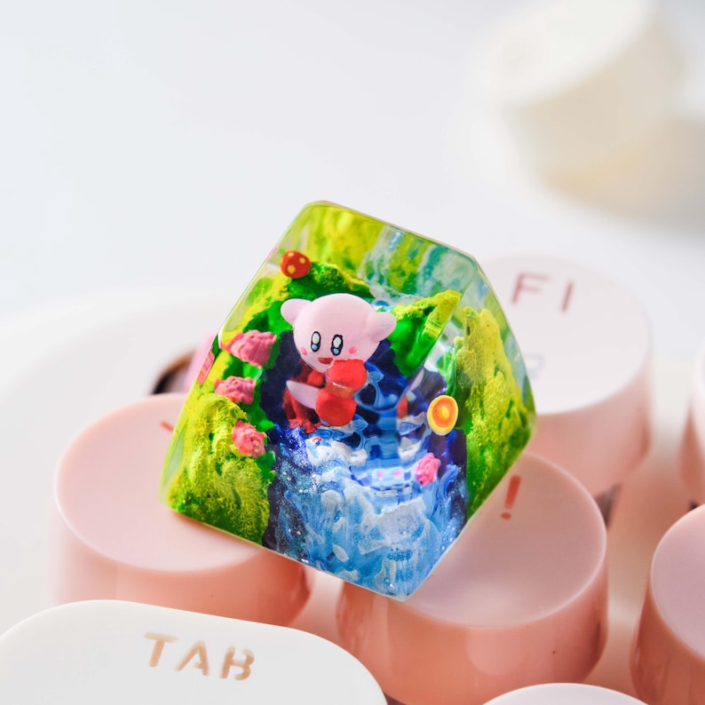 May include: A translucent resin mechanical keyboard keycap featuring a miniature pink character with red accents, set within a landscape of green hills, blue water, and small red mushrooms, resting on top of light pink keyboard keys.