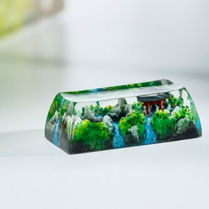 May include: A custom keycap featuring a miniature landscape scene. The keycap is clear, encapsulating a detailed diorama of a waterfall, green foliage, gray rocks, and a small red structure. The keycap is rectangular with a curved top.