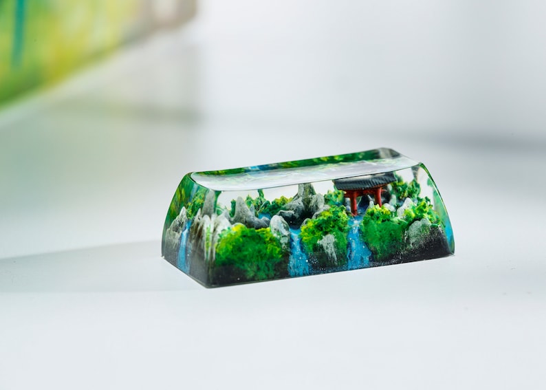 May include: A custom keycap featuring a miniature landscape scene. The keycap is clear, encapsulating a detailed diorama of a waterfall, green foliage, gray rocks, and a small red structure. The keycap is rectangular with a curved top.